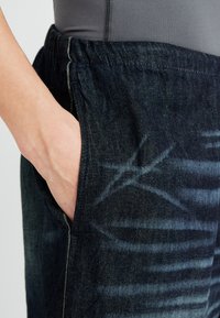 Denimhousut joustavalla vyötärönauhalla, tummansininen väri, jossa on sivutasku ja hillitty pesuainekuvio, joka tuo tekstuuria ja yksityiskohtia.