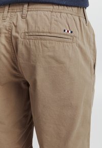 Beige bomullsbukser med en baklomme med broderte tri-farge aksenter. Tøyet har en myk, glatt tekstur og en avslappet passform.
