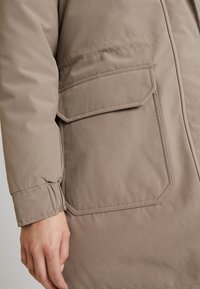 Ruskea, toppaustäytteinen ulkotakki, jossa lähikuva taskusta ja teksturoidusta kankaasta. Takissa on resorit hihansuissa ja näkyvät saumat.