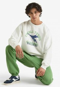 Felpa bianca con grafica "Circolo Tennis" in verde e blu, abbinata a pantaloni della tuta verdi. Sneakers blu navy completano l'outfit.