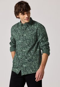 Zelená košile na knoflíky s motivem listů, dlouhé rukávy ohrnuté, černé knoflíky a rovný lem. Nosená s černými kalhotami.