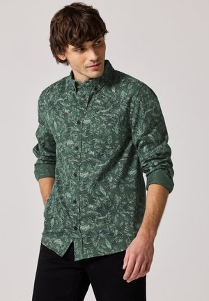 Groene overhemdd met een bladpatroon, lange mouwen opgerold, zwarte knopen en een rechte zoom. Draagt bij zwarte broek.