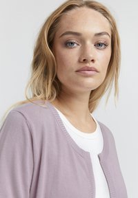 Gilet lilas avec une texture tricot légère, encolure ronde et un design ouvert, superposé à un t-shirt blanc.