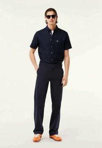 Camisa de botão de manga curta azul-marinho com um pequeno logotipo, combinada com calças azul-marinho escuras e ténis laranja contra um fundo simples.