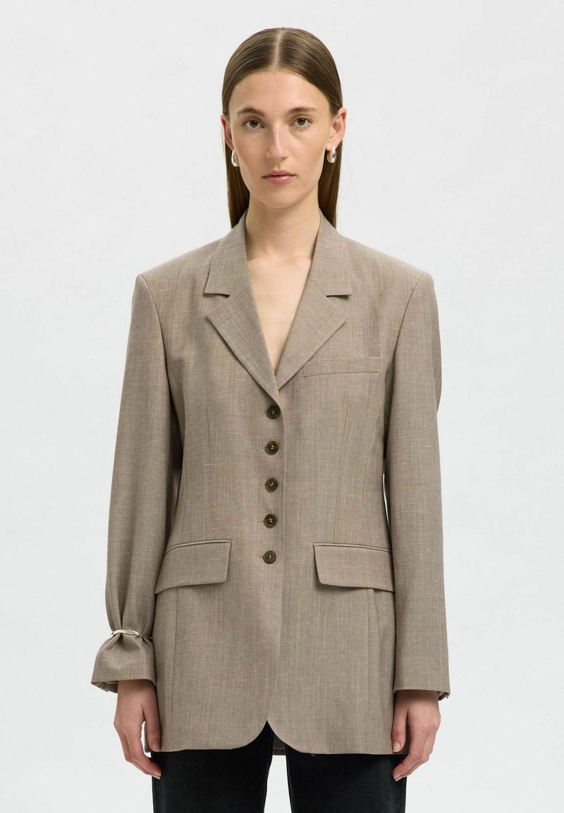 Mujer con cabello castaño lacio viste una chaqueta oversized color marrón claro con cinco botones y bolsillos con solapa, combinada con pantalones negros, fondo neutro.