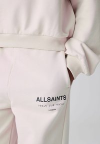Jasnobeżowe spodnie dresowe z kieszenią, w której znajduje się ręka, z czarnym napisem "ALLSAINTS" oraz japońskimi znakami nad "LONDYN" na nogawce.