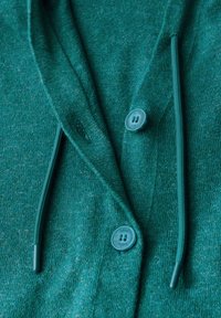 Teal gebreide cardigan met een gestructureerde afwerking, voorzien van een kraag, twee knopen en koorden. Het materiaal is zacht en iets dik.