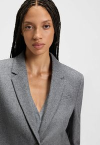 Giovane donna con capelli intrecciati che indossa un blazer grigio sopra un top grigio a scollo a V, che guarda con calma la fotocamera su uno sfondo neutro.