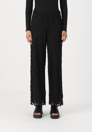 Black wide-leg pants made of textured lace fabric, featuring a scalloped hem and drawstring waist, paired with black platform sandals.