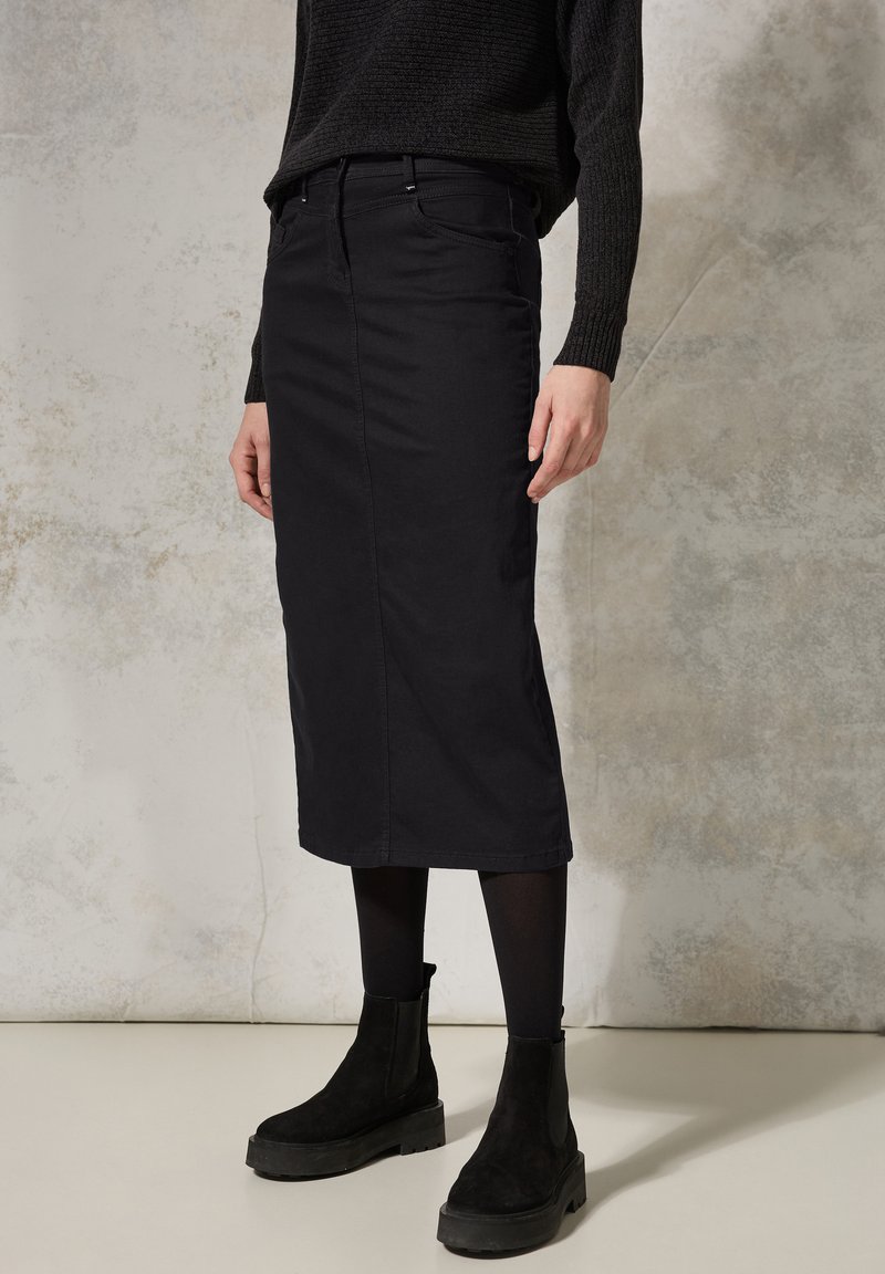 Falda midi de mezcla de algodón negra con corte recto, bolsillos frontales y sutiles detalles de costura, combinada con botas negras hasta el tobillo y medias.
