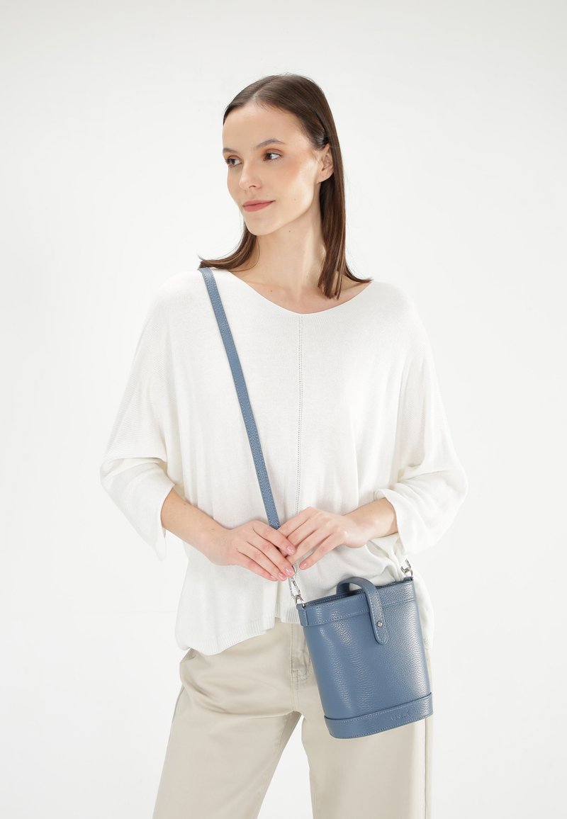 Lichtblauwe leren emmertas met een gestructureerde afwerking, korte handgreep en verstelbare schouderriem, gecombineerd met een witte top en beige broek.
