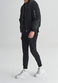Persona vistiendo una chaqueta bomber negra, pantalones negros, calcetines blancos y zapatillas blancas de pie contra un fondo claro y liso.