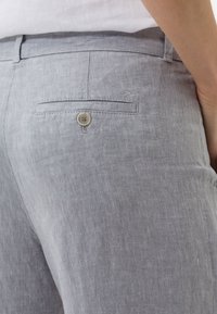 Šedé lněné kalhoty s zadní kapsou na knoflík a jemnými švy. Textura se zdá být lehká a prodyšná.