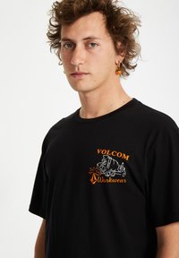 T-shirt en coton noir avec un grand graphique orange et blanc sur la poitrine, comportant le texte "VOLCOM Workwear" et un motif de mélangeur à béton.