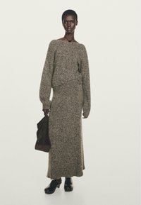 Modella che indossa un abito coordinato composto da un lungo maglione di maglia e una gonna grigia, tiene in mano una borsa in suede marrone e indossa ciabatte nere con suola in legno.