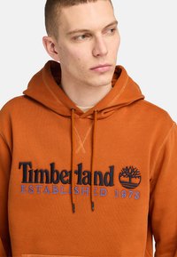 Pomarańczowy dres z kapturem wykonany z miękkiego materiału, z czarnym i niebieskim logo Timberland oraz grafiką drzewa, posiadający ściągacze.