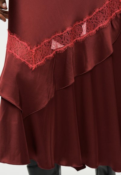 Dieprode satijnen rok met gerafelde zoom en diagonale kanten bies, gedragen met zwarte laarzen, hand die de stof aan de linkerkant aanpast.