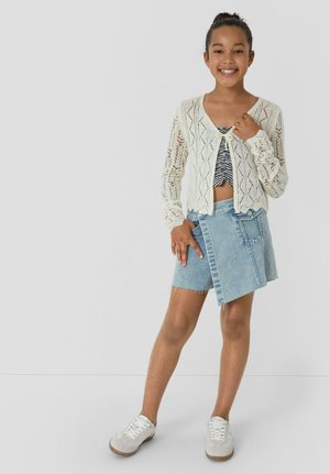 Lächelndes Mädchen, das eine weiße Strickjacke, ein bauchfreies Zebra-Print-Oberteil, einen asymmetrischen Jeansrock und weiße Sneakers trägt, vor einem schlichten Hintergrund steht.