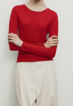 Person wearing a fitted red long-sleeve top and high-waisted, loose beige pants, standing with arms crossed against a plain background.