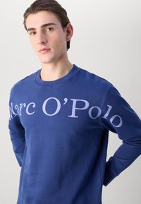 Sudadera de cuello redondo en azul marino, hecha de una mezcla de algodón, con un gran estampado de logo en color lila claro en el pecho. Textura suave y corte casual.