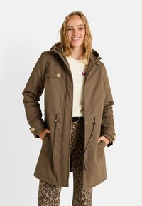 Abrigo marrón hasta la rodilla con capucha, con detalles de botones, cintura ajustable con cordón y dos bolsillos delanteros, llevado sobre un suéter ligero y pantalones estampados.