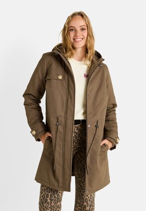 Abrigo marrón hasta la rodilla con capucha, con detalles de botones, cintura ajustable con cordón y dos bolsillos delanteros, llevado sobre un suéter ligero y pantalones estampados.