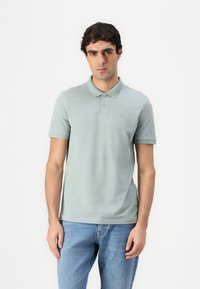 Lys grønn polo shirt med krage, tre knapper og et brodert logo. Kombinert med lyseblå denim jeans, båret på en avslappet måte.