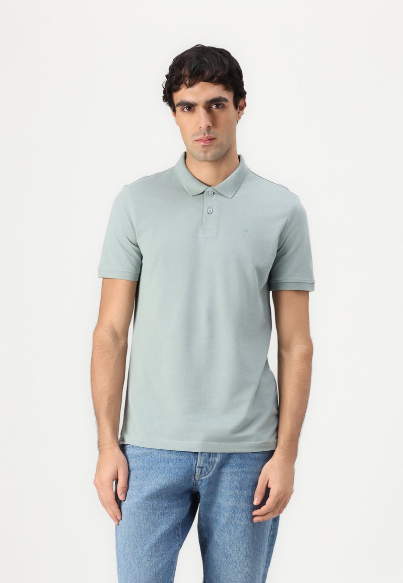 Lys grønn polo shirt med krage, tre knapper og et brodert logo. Kombinert med lyseblå denim jeans, båret på en avslappet måte.