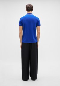 Kék pólóing gallérral, rövid ujjú, egyenes szegéllyel. Laza, fekete, széles nadrággal párosítva. Simább textúra, minimalista dizájn.