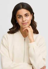 Cream-colored bomber jacket with a textured finish, ribbed cuffs, and a front zipper closure. Model is resting chin on hand.