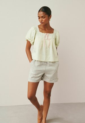 Junge Frau trägt eine hellgrüne Bluse mit Puffärmeln, pinker Krawatte und beige karierte Shorts, steht mit den Händen in den Taschen vor einem einfarbigen Hintergrund.