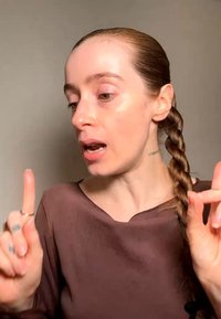 Vrouw met vlechten en een bruin topje die spreekt en gebaart met opgestoken wijsvingers tegen een effen achtergrond.