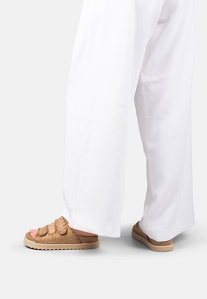 Piernas con pantalones blancos holgados y sandalias abiertas color cuero con tiras texturizadas y suelas gruesas sobre un fondo blanco liso.