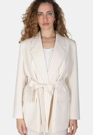 Donna con capelli ricci indossa un blazer beige a righe sottili legato in vita sopra una maglietta bianca, in piedi davanti a uno sfondo semplice.