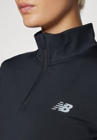 Pullover nero a mezzo zip realizzato in tessuto morbido, presenta un colletto alto e un logo grigio riflettente sul petto.