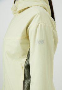 Chaqueta deportiva amarilla clara con capucha, que presenta un interior texturizado verde, bolsillos con cremallera y un logotipo impreso en la manga.