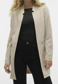Béžový texturovaný blazer s jednobuttonovým zapínáním, dvěma zipovými kapsami a dlouhým, rovně střiženým siluetou, nošený přes černý top a kalhoty.