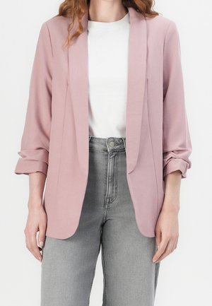 Vrouw draagt een lichtroze blazer over een wit topje, gecombineerd met hooggetailleerde grijze jeans tegen een effen lichte achtergrond.