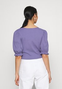 Top corto de lavanda con mangas abullonadas y puños recogidos. Textura suave y escote redondo, combinado con pantalones blancos de tiro alto.