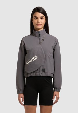 Woman wearing a gray zip-up jacket with a matching crossbody bag labeled "KHUJO 2001" and black cycling shorts, standing against a gray background.