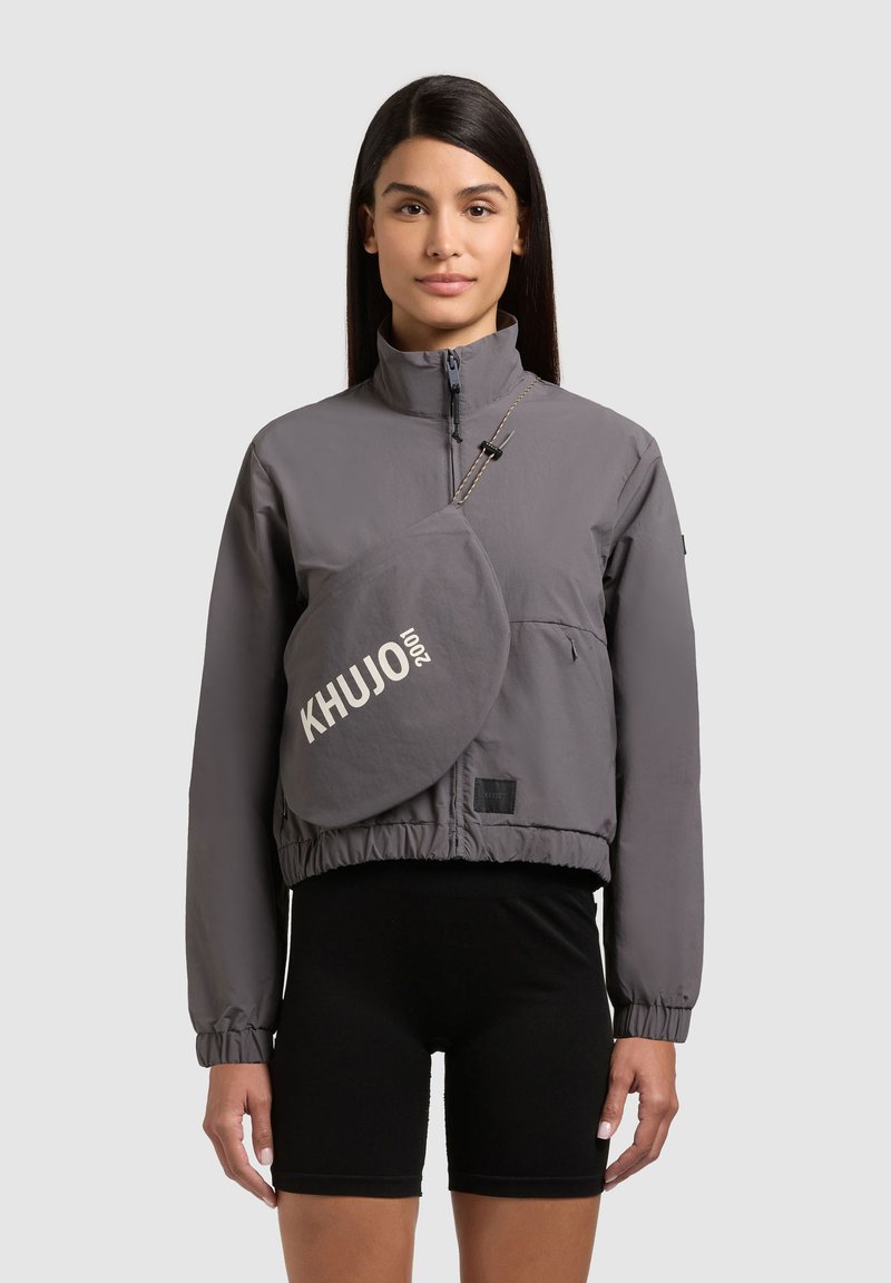 Femme portant une veste grise zippée avec un sac bandoulière assorti portant l'inscription « KHUJO 2001 » et un short de cyclisme noir, debout devant un fond gris.
