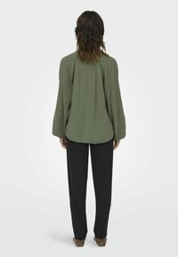 Olivgrüne, strukturierte Bluse mit Ballonärmeln, getragen über schwarzen Hosen. Die Bluse hat eine lockere Passform und einen abgerundeten Saum.