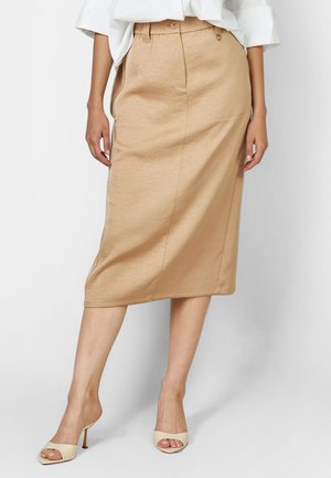 Woman wearing a beige midi pencil skirt with a button waist and white high-heeled open-toe sandals, standing against a white background.