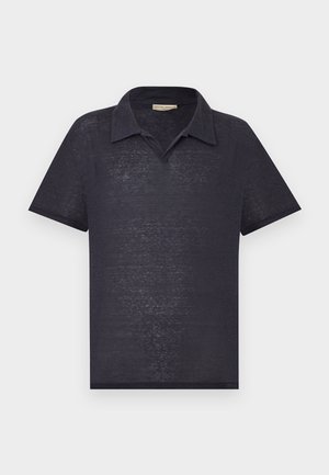 Άνδρας με κατσαρά μαλλιά, φορώντας μαύρο πόλο με κοντά μανίκια, γκρι φαρδιά παντελόνια και μαύρα παπούτσια, στέκεται με τα χέρια στις τσέπες.