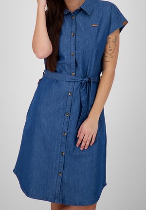 Femme portant une robe chemise en denim bleu avec une ceinture à la taille, des manches courtes et un tatouage minimaliste sur le visage de son bras gauche.