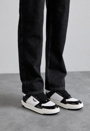 Persona que lleva zapatillas bajas negras y blancas con cordones negros y vaqueros negros, de pie sobre un suelo gris.