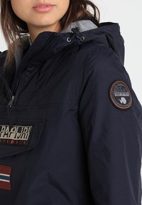 Veste bleu marine avec une demi-fermeture éclair, capuche grise et patchs de logo. Dispose d'une poche avant et d'un tissu texturé. Matériau lisse et léger.