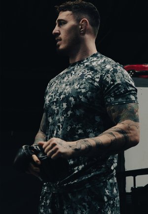 Camouflage-patterned athletic shirt made of breathable fabric; holds black boxing gloves. Close up on arms with detailed tattoos.
