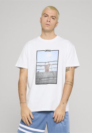 Giovane uomo con capelli biondi corti che indossa una maglietta bianca con una grafica "UNFAIR" e pantaloncini blu con strisce bianche.