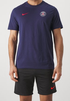 Homme portant un t-shirt bleu marine Paris Saint-Germain avec un logo Nike rouge et un short noir avec un logo Nike rouge, debout devant un fond uni.